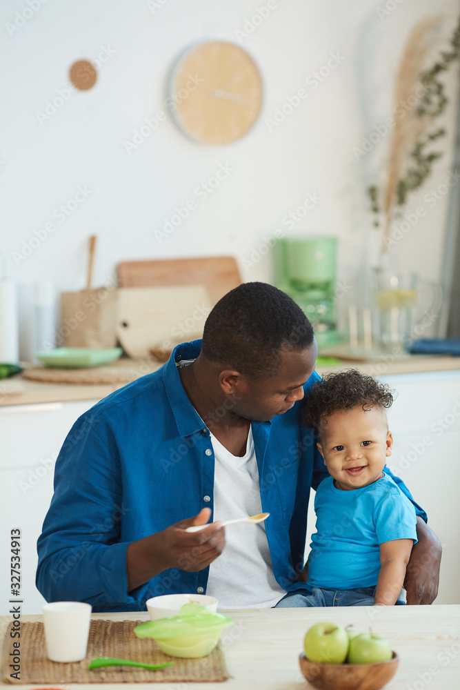 Portrait of happy African-American dad feeding cute little boy smiling at camera white sitting at kitchen table, copy space Portrait of happy African-American dad feeding cute little boy smiling at camera white sitting at kitchen table, copy space