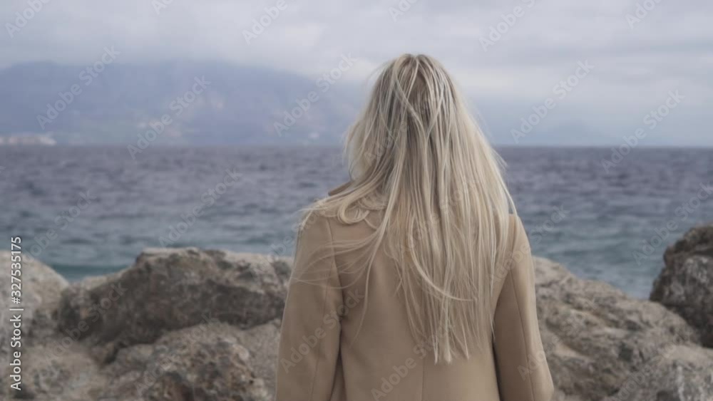 A girl with blond hair in a beige coat walks along a rocky seashore A girl with blond hair in a beige coat walks along a rocky seashore