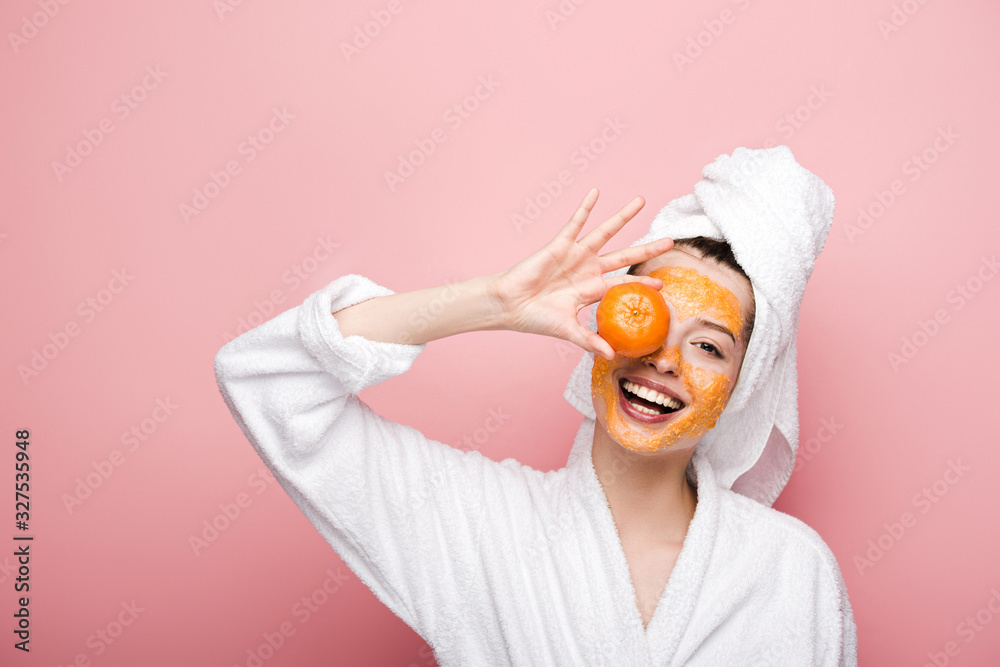 cheerful girl with citrus facial mask covering eye with tangerine on pink background