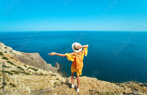 Back view of woman traveler in bright yellow dress and hat with backpack standing the top of mountain with raised hands against amazing seascape. © vitaliymateha