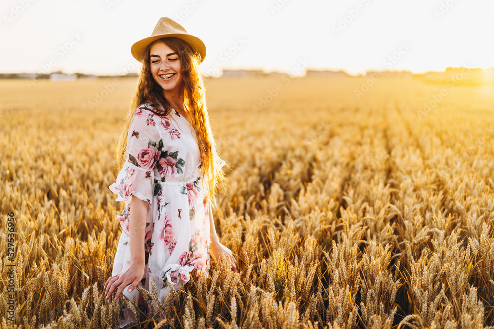 Portrait of a beautiful young woman with curly hair and freckles face. Woman in dress and hat posing in wheat field at sunset and looking at camera