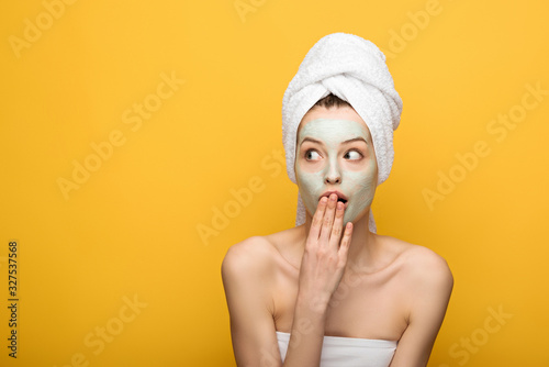 shocked girl with nourishing facial mask covering mouth with hand and looking away isolated on yellow
