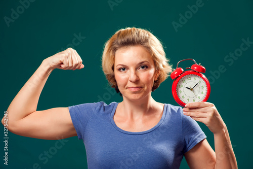 A portrait of smiling woman holding alarm clock and showing her bicep. People and emotions concept