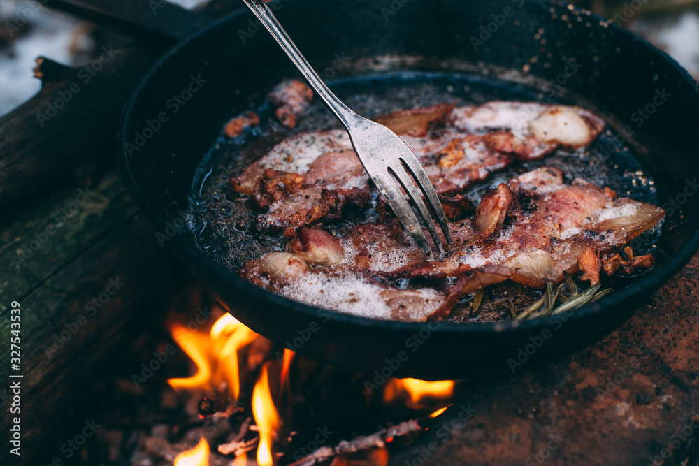 Slices of fried bacon in a pan. Food in a forest camp. Cooking on fire. Picnic in the nature. Grilled food on nature. Fork taking a slice of bacon.. Slices of fried bacon in a pan. Food in a forest camp. Cooking on fire. Picnic in the nature. Grilled food on nature. Fork taking a slice of bacon..