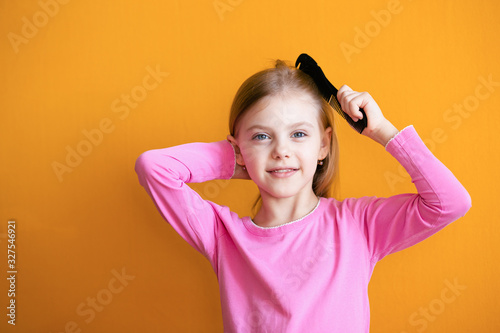Cute baby, baby girl 6-8 years old combs her soft blond hair medium length comb and smiles on an orange background