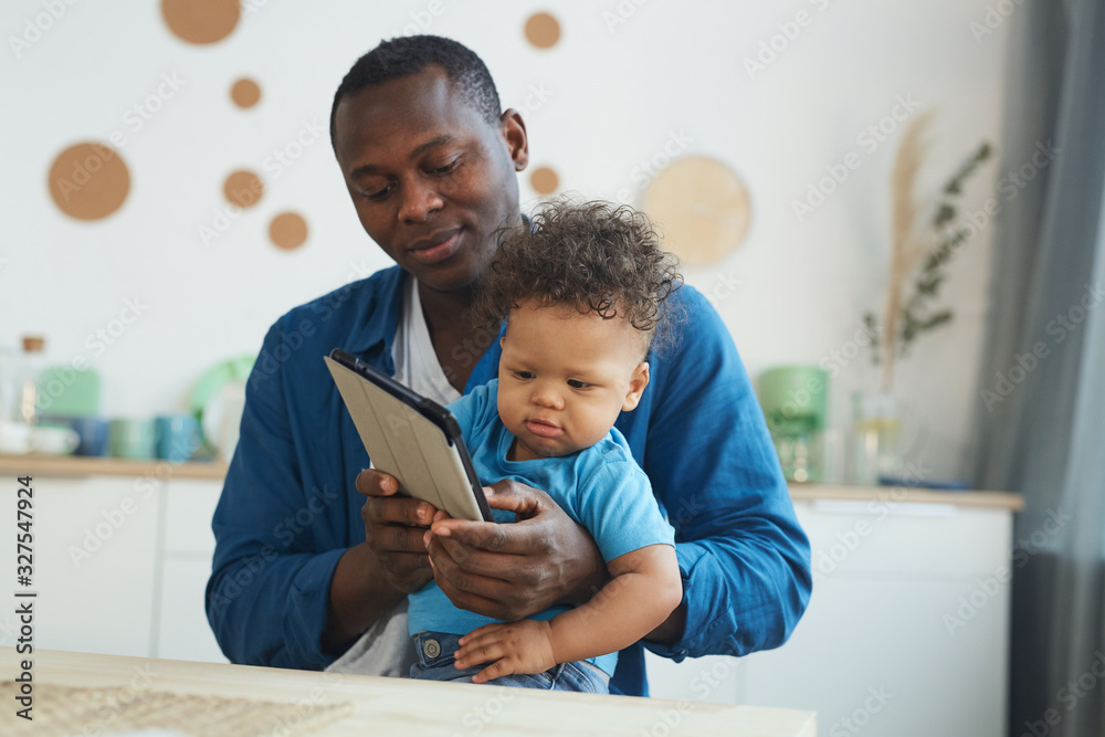 Portrait of African-American man holding crying baby while using digital tablet in home interior, copy space Portrait of African-American man holding crying baby while using digital tablet in home interior, copy space
