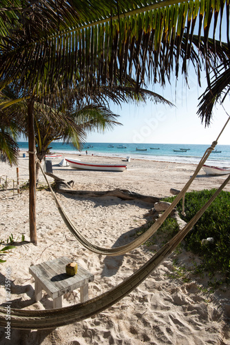 Fototapeta Naklejka Na Ścianę i Meble -  Hammocks on a Caribbean beach in the shade of palm trees. In the background, fishing boats on a beach in Tulum Mexico