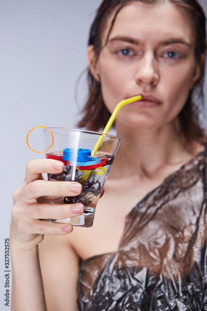 The environmental pollution is destroying human health and our planet. A glooming lady in a black polythene wrapper is posing with a glass with a water, polythene trash and a yellow straw. The environmental pollution is destroying human health and our planet. A glooming lady in a black polythene wrapper is posing with a glass with a water, polythene trash and a yellow straw.