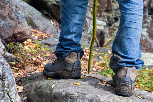 Legs of a tourist in hiking boots on a walk in the autumn along a mountain trail. A man in black boots and blue jeans against a background of mountain boulders. Vacation and adventure concept.