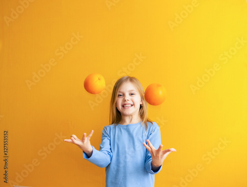 Cute baby 6-8 years old, baby girl blonde beauty on an orange background throws and catches fruits appelsinandand laughs,