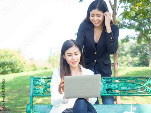 two asia business woman working with computer laptop in park. beauty woman tpying keyboard notebook blank screen and friend standing behind with copy space.   photo