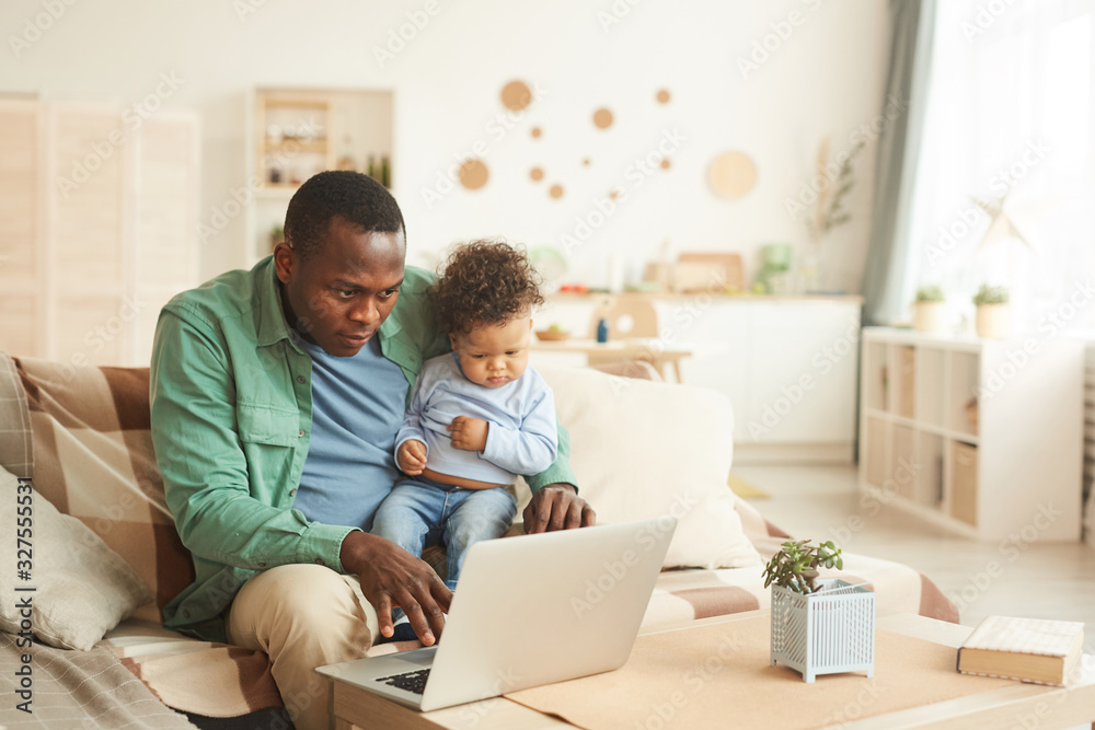 Portrait of mature African-American man working on laptop while babysitting son at home, copy space Portrait of mature African-American man working on laptop while babysitting son at home, copy space
