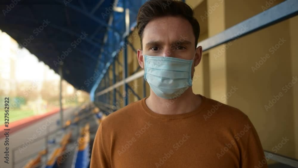 Man in surgical mask standing on empty stadium. Man puts the mask on of his face while pointing finger to mouth, coughing. afraid of coronavirus wearing mask to protect from epidemic virus disease. Man in surgical mask standing on empty stadium. Man puts the mask on of his face while pointing finger to mouth, coughing. afraid of coronavirus wearing mask to protect from epidemic virus disease.