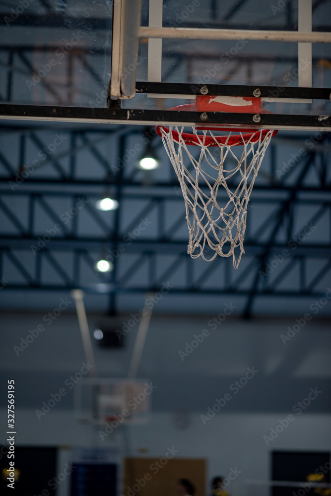 An interior space of a basketball court showing a scaffolding metal roof sheet with a framework of metal truss with sunlight goes through and a basketball ring as a focal point.