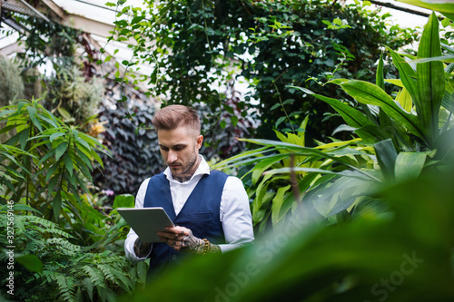 Young man with tablet standing in botanical garden, working.