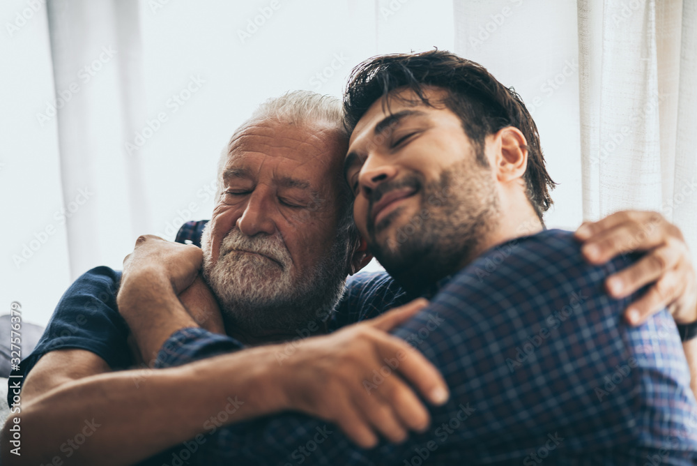 Sweet bonding. Cheerful elderly man sitting on the sofa next to his adult son, hugging him and posing together with him