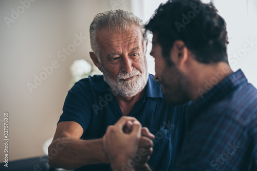 Sweet bonding. Cheerful elderly man sitting on the sofa next to his adult son, hugging him and posing together with him, concept of Father's Day