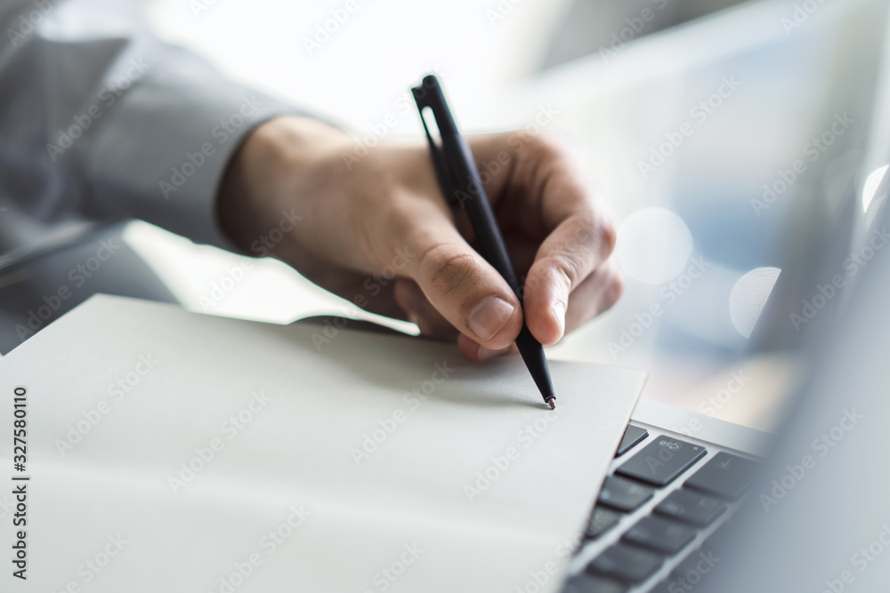 Businessman writes with a pen in diary on laptop keyboard in a sunny office, business and education concept. Close up