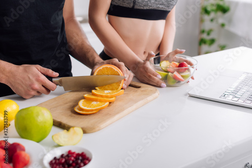 cropped view of man cutting orange near woman, salad and laptop
