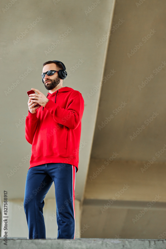 Young attractive bearded man standing under the bridge, listening to the music and using phone for texting messages.