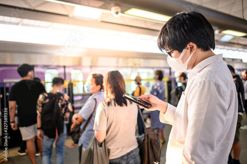 Asian man wearing surgical face mask using smartphone at skytrain station platform. Wuhan coronavirus (COVID-19) outbreak prevention in public transportation. Health awareness for pandemic protection