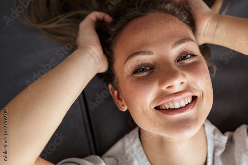 Image of happy caucasian woman smiling at camera while lying on couch