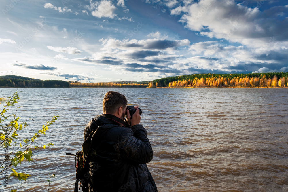 Rear view of the figure of a photographer taking a picture on the shore of a forest lake in autumn.