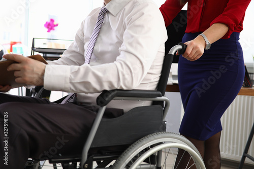 Woman carries man in suit on wheelchair workplace. Person moves on wheelchair. Interview invitation. Wheelchair access to office. Person with disabilities returned to active work workplace