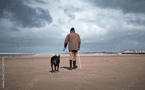 A retired old man with a walking stick walking his dog on a beach. Dogs are mans best friend and a loyal companion for older people who are alone. Love animals and pets.