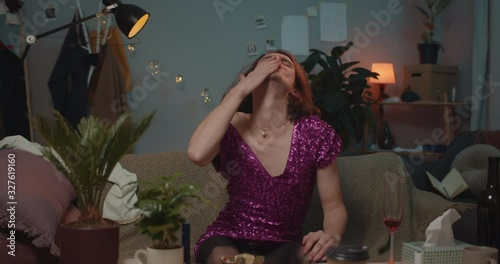 Drunky transgender man in fashionable dress with sequins wiping lipstick and grabbing head. Upset transgender sitting on sofa after broke up with boyfriend. Concept of emotions and feelings. photo