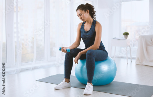 Latin woman in sportswear working out with dumbbells and fitness ball indoors, copy space text