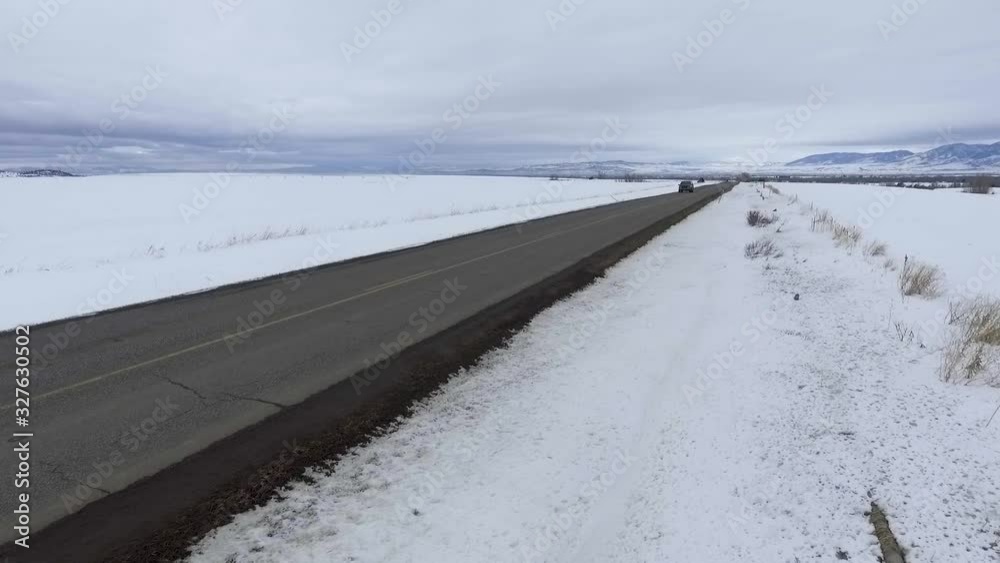 Looking down the long snow surrounded road, a blue suburban quickly approaches and the drone pans to follow it Looking down the long snow surrounded road, a blue suburban quickly approaches and the drone pans to follow it