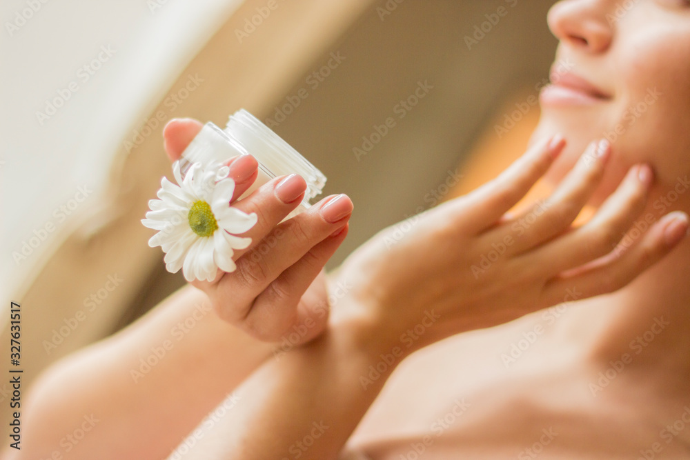 Spring portrait of gentle pretty girl looking away and smiling while applying moisturizing lotion on face. Beautiful young woman holding jar of cream in hand. Gentle girl, skin or body care, treatment