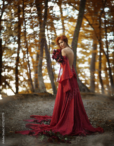 Amazing cute young woman with fiery red hair in beautiful long red dress with an open back holds large bouquet of roses standing with her back to camera on path with roses in yellow autumn forest