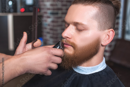 A male barber at work, he does a beard and mustache haircut to a man in the barbershop. Male cosmetology, care of the beard, creating a stylish look.