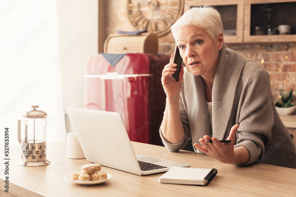 Concerned elderly woman talking on cellphone, working on laptop in kitchen Concerned elderly woman talking on cellphone, working on laptop in kitchen