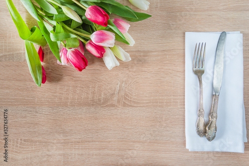 Romantic table setting. Vintage fork and knife on white napkin and bunch of purple and white tulips on wooden background. Top view, space for text