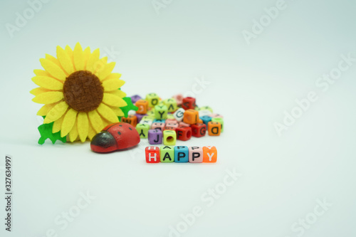 Alphabet beads arranged in the word "HAPPY" and a pile of different colored beads, paper sunflower, wooden red ladybug are on the back, placed on a white background.