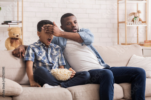 Father and son watching scary movie together, dad covering kid's eyes