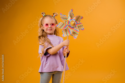 little blonde girl smiling in sunglasses holding a windmill stands on a yellow background in the Studio isolate. happy child, text space, horizontal photo