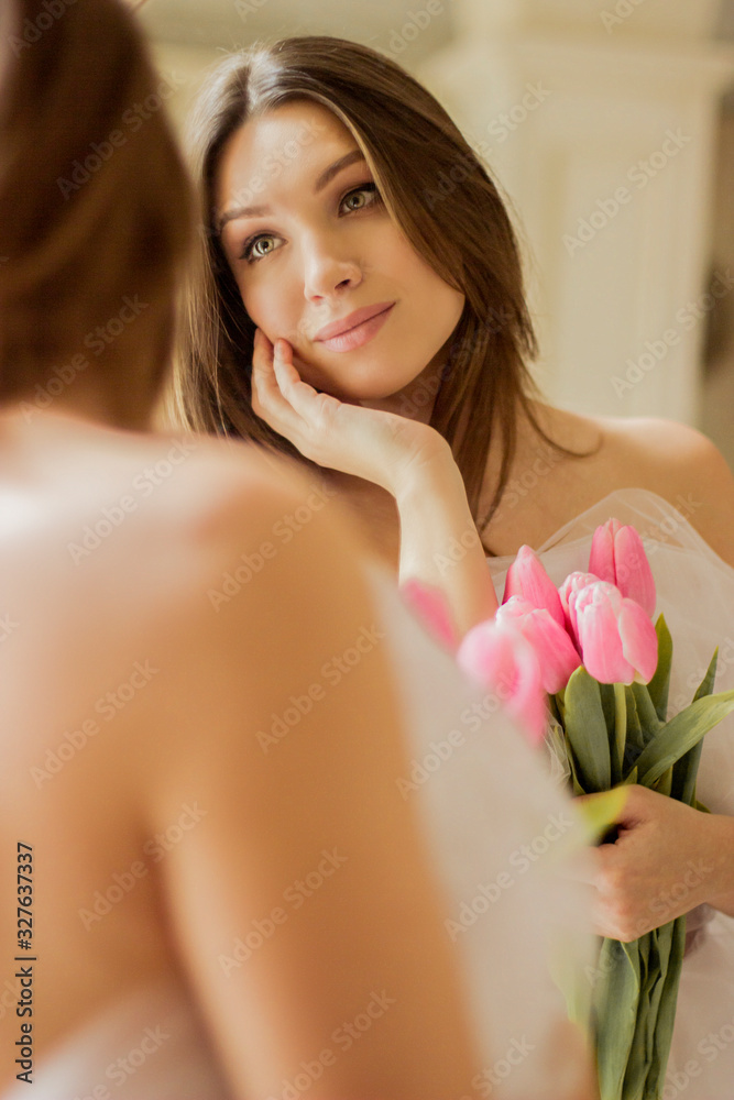 Closeup of young beautiful woman looking at her reflection in mirror and holding bouquet of spring flowers. Pretty and tender girl in white dress posing against mirror with tulips in hands. 8 march Closeup of young beautiful woman looking at her reflection in mirror and holding bouquet of spring flowers. Pretty and tender girl in white dress posing against mirror with tulips in hands. 8 march