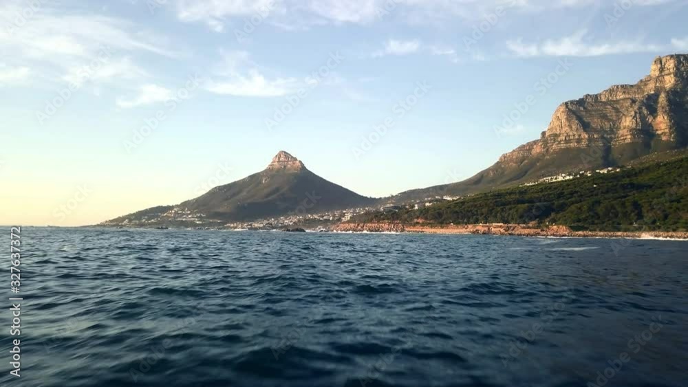 Camps Bay during early sunset in a romantic evening golden hour light. Wide reveal aerial shot showing ocean, houses and Lions Head mountain. Drone flying on low altitude over ocean towards coast line Camps Bay during early sunset in a romantic evening golden hour light. Wide reveal aerial shot showing ocean, houses and Lions Head mountain. Drone flying on low altitude over ocean towards coast line