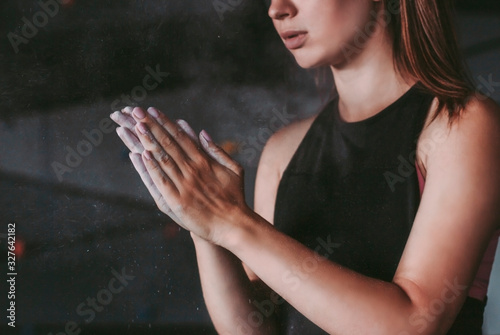 Closeup of young attractive woman athlete clapping hands with workout dust while standing in dark gym. Beautiful determined sports girl with gym chalc on arms. Preparation for exercising with barbell photo