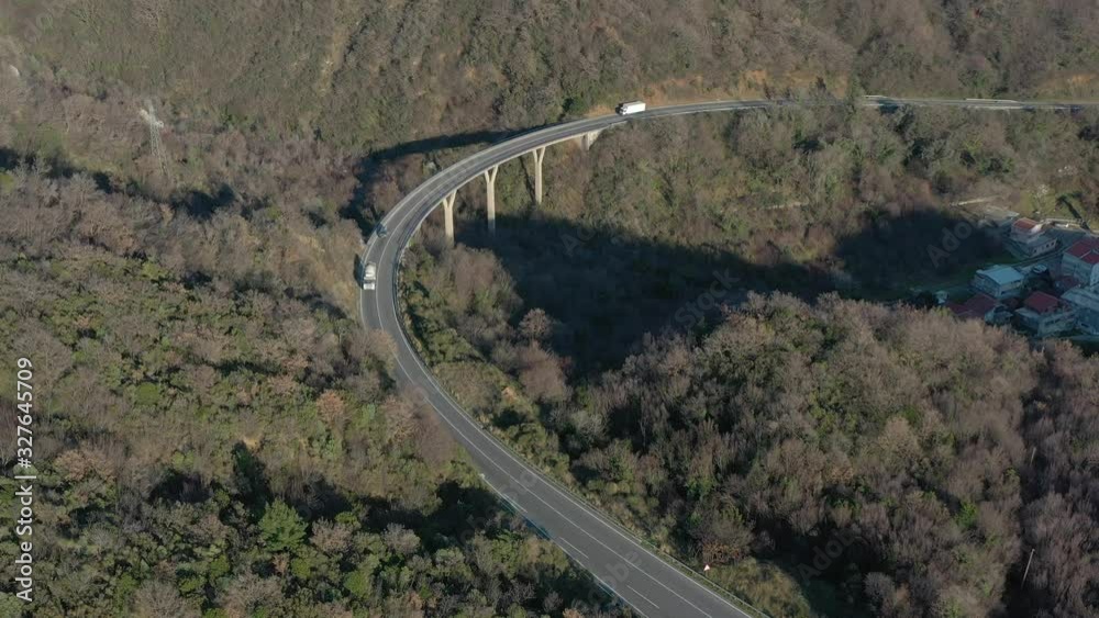 Aerial shot of a narrow winding mountain road, a bridge on which cars and trucks go. Around mountains covered with coniferous forest are seen Aerial shot of a narrow winding mountain road, a bridge on which cars and trucks go. Around mountains covered with coniferous forest are seen