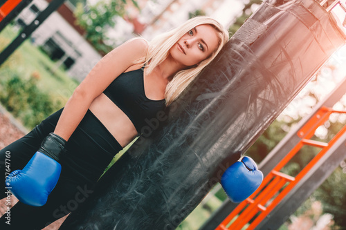 Portrait beautiful blonde fitness girl in boxing gloves hugging punching bag after training outdoors. Young attractive sports woman posing with boxing bag on sports ground outdoors. Female martial art