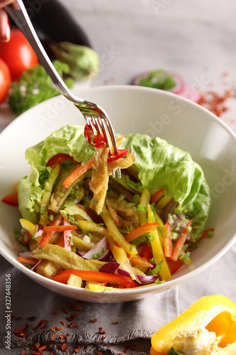Healthy food. Vegetable salad in a deep light bowl. Tomatoes, peppers, onions, lettuce, eggplant, fork and spices on a light grey background. Background image, copy space