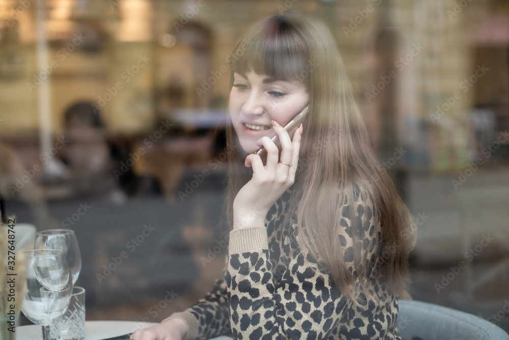 Fototapeta premium Young beautiful girl outside the window in a cafe talking on a mobile phone.
