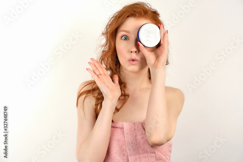 A red-haired curly girl in a pink towel stands on a white background, covering eye with a jar of cream and stretching lips forward