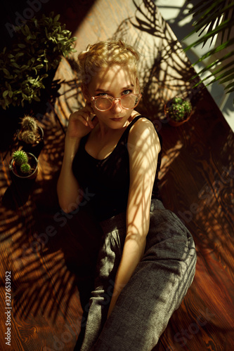 Top view of beautiful woman in retro glasses, posing on wooden background surrounded by cactuses and palms. Nature interior design. Sunlight and plant shadows.