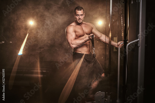 Athletic shirtless sportsman doing an excersise on a hand pull machine in a dark gym under the spotlights surrounded by smoke